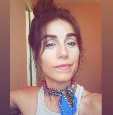 Uma mulher de cabelos escuros amarrados usa um lenço colorido no pescoço. Ela está dentro de casa, olhando confiante para a câmera com um leve sorriso.