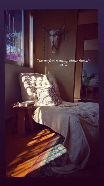 Uma cadeira de leitura aconchegante e iluminada pelo sol fica ao lado de uma janela, coberta com um cobertor. Um travesseiro, uma mesa lateral com livros e uma caveira decorativa criam um recanto tranquilo.