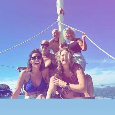 Fünf Freunde lächeln fröhlich auf einem Boot unter sonnigem Himmel. Sie tragen Badeanzüge und Sommerkleidung und genießen einen lebhaften Tag auf dem Meer mit blauem Wasser und Himmel.