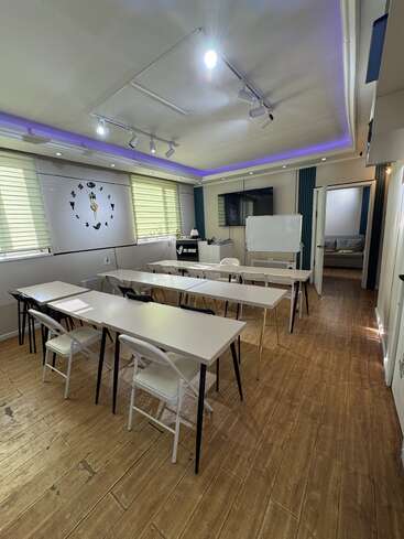 This image shows a modern classroom with long tables, folding chairs, a large wall clock, a whiteboard, TV screen, and wooden-style flooring with overhead lighting.