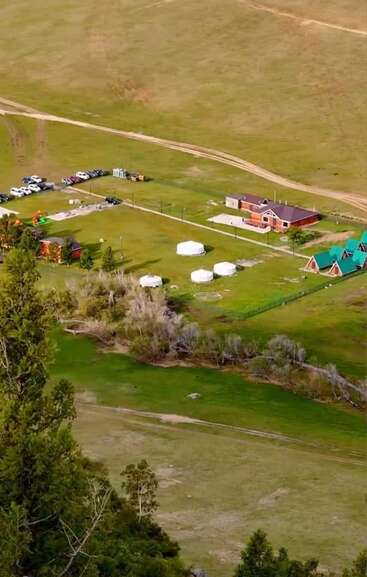 This aerial image shows a rural property with several buildings, traditional yurts, parked cars, green lawns, and trees, surrounded by open fields and dirt roads.