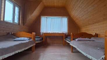 This cozy attic room features wooden walls, three single beds with blankets, pillows, a window with sheer curtains, and a small wooden table beneath the window.