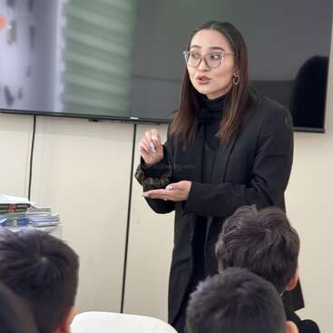 A woman, likely a teacher, stands in front of a classroom holding a marker and speaking to a group of attentive students. Books are stacked nearby.