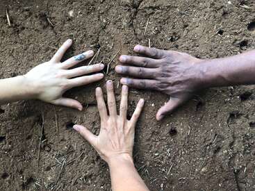 Three hands of different skin tones are spread open on brown soil, symbolizing unity, diversity, and connection with nature. Small holes are visible in the ground.