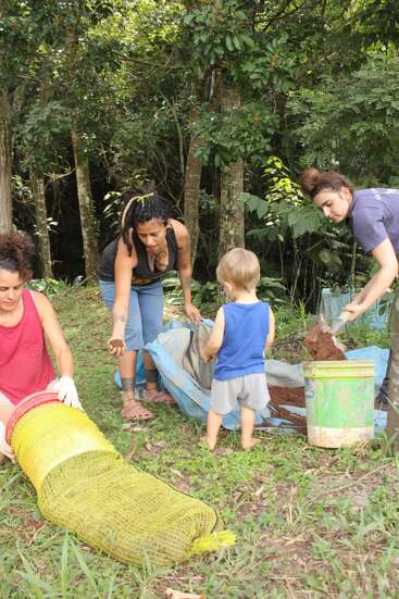 Four people, including a child, are outdoors working together with soil and sacks. They appear to be participating in a gardening or environmental project surrounded by trees and greenery.