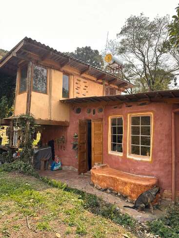 This rustic, two-story house features earthy tones, large windows, and natural materials. A dog rests by the entrance, and greenery surrounds the cozy, inviting home.