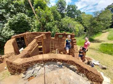 Drei Leute stehen in einem noch nicht fertig gebauten Lehmhaus, das von viel Grün umgeben ist. Das Haus ist aus natürlichen Materialien gebaut und zeigt, wie man nachhaltig in einem Wald bauen kann.
