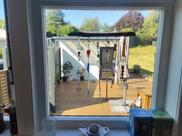 Through a window, a decorative wind chime hangs. Outside is a sunny wooden deck with potted plants, seating, umbrella, and a grassy backyard garden.