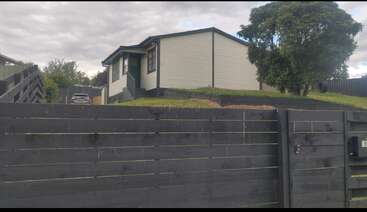 A small, modern house sits atop a grassy hill, surrounded by a tall black wooden fence and a large tree, with a car parked nearby.
