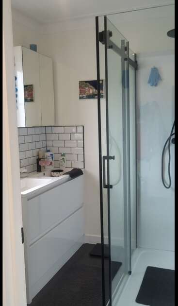 A modern bathroom featuring a glass shower, white tiles, a sleek vanity, wall cabinet, black hardware, dark floor mats, and small decor accents throughout.