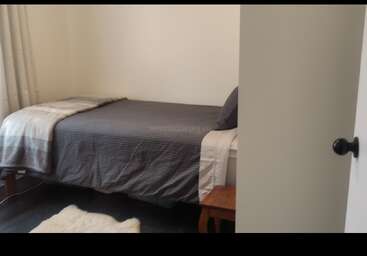 A small, simple bedroom featuring a single bed with gray bedding, a wooden nightstand, a white rug, neutral walls, and a partially open door.