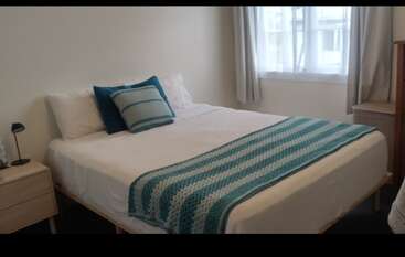 A cozy bedroom features a neatly made bed with white linens, blue-striped pillows, and a matching blanket. Natural light streams through the curtained window, creating a relaxing atmosphere.