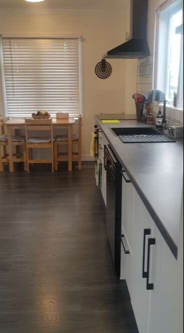 This image shows a modern kitchen with dark flooring, a long counter, white cabinets, sink, utensils, window, dining table with chairs, and blinds covering the window.
