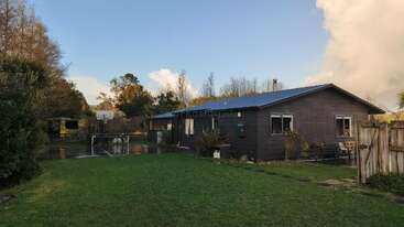 A cozy single-story house with a blue roof sits amid green grass and trees. There’s a basketball hoop and court, patio furniture, and a wooden fence.