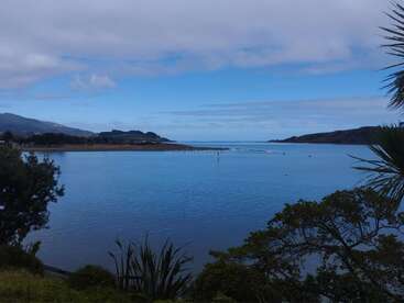 A tranquil bay with calm blue waters, surrounded by lush green trees and distant hills under a partly cloudy sky, creating a peaceful natural landscape scene.