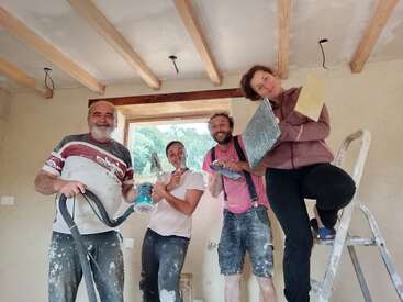 Four people, smiling and posing with construction tools inside a house under renovation. Paint on their clothes suggests teamwork, creativity, and effort. One stands on a ladder.