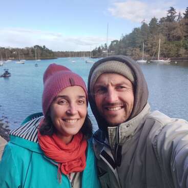 A smiling couple dressed warmly pose for a selfie by a scenic harbor, with calm blue water, sailboats, and tree-lined shore under a partly cloudy sky.
