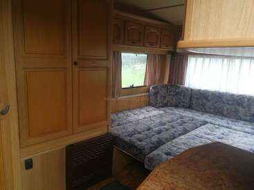 L'image représente l'intérieur d'un camping-car, avec un canapé, des armoires et une fenêtre, avec une table en bois au premier plan et un champ herbeux à l'extérieur.