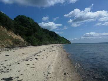 L'image représente une scène de plage sereine avec un rivage sablonneux, une colline escarpée couverte de verdure et une étendue d'eau sous un ciel bleu avec des nuages blancs.