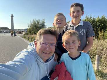 A man and three boys smile outdoors for a selfie. The sun is shining, there is greenery, and a tall lighthouse stands in the background. Happy moment.