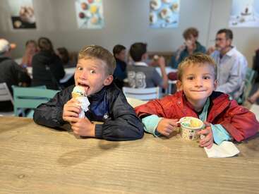 Dos niños disfrutan de un helado en una mesa de madera de una concurrida heladería, rodeados de otras personas. Ambos parecen felices y emocionados.