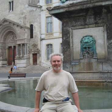 Um cara de cabelo grisalho tá sentado na beirada de uma fonte com uma cara de pedra. Atrás dele, tem uma igreja europeia histórica com uma arquitetura bem ornamentada.