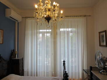 Cette photo montre une chambre à coucher classique avec des rideaux blancs vaporeux, un lustre richement décoré, des meubles en bois, un climatiseur, un ventilateur et des tableaux accrochés aux murs. L'ambiance est lumineuse.