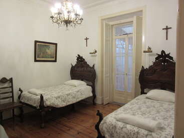Cette chambre comprend deux lits en bois richement décorés, un lustre classique, une chaise en bois, des croix religieuses, des tableaux encadrés, de la literie blanche et de grandes portes-fenêtres. Ambiance chaleureuse.