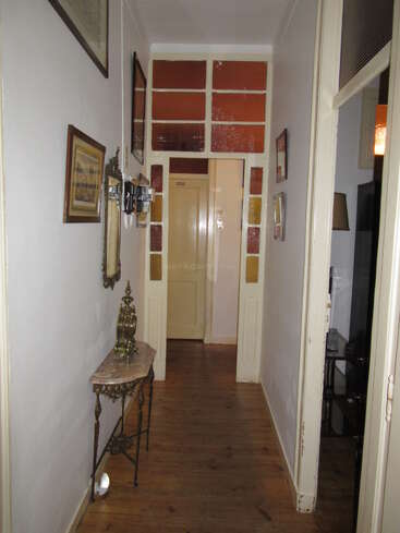 Un couloir étroit au sol en parquet, orné de photos encadrées, d'un miroir et d'une table vintage sur laquelle trône une horloge décorée, mène à une porte beige.