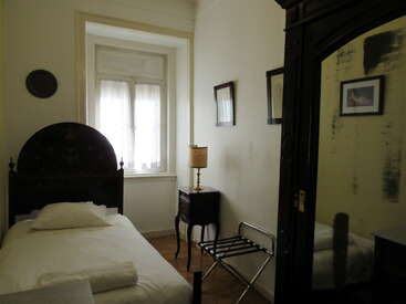 Une petite chambre confortable comprend un lit simple, une tête de lit vintage, une table de chevet avec une lampe, une armoire à miroirs, des photos encadrées, un porte-bagages et une fenêtre baignée de soleil.