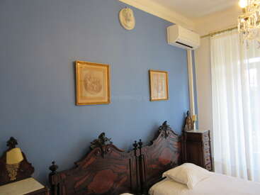 Cette élégante chambre à coucher est équipée de lits jumeaux en bois sculpté, d'un mur bleu, de tableaux encadrés, de tables de chevet classiques, d'un lustre, d'un climatiseur et de grandes fenêtres ornées de voilages.