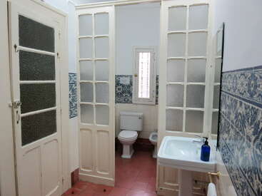 Il s'agit d'une salle de bains avec un sol en carrelage rouge, un lavabo blanc, des carreaux à motifs bleus, des portes en verre dépoli, des toilettes, un bidet, une petite fenêtre et un miroir.