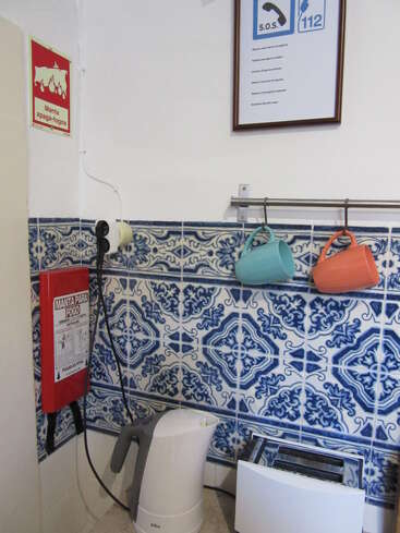 Dans un coin cuisine orné de carreaux bleus et blancs à motifs, on trouve des tasses suspendues, une bouilloire électrique, un grille-pain et une couverture anti-feu fixée au mur pour des raisons de sécurité.