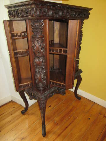 Il s'agit d'un meuble ancien en bois richement décoré, orné de sculptures complexes, doté de portes ouvertes, de pieds courbés et de plusieurs étagères. Il se dresse sur un parquet ciré, devant un mur jaune.