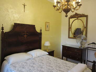 Cette image montre une chambre à coucher classique avec un lit en bois richement décoré, un élégant lustre, un grand miroir, une table de chevet, un ventilateur, un crucifix et une commode de style vintage.