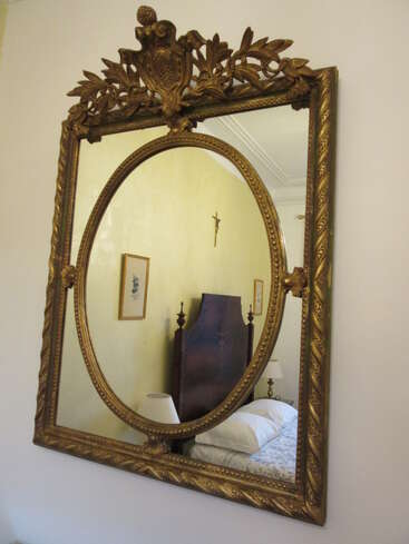 Un miroir mural orné d'un cadre doré reflète une chambre à coucher classique, avec une haute tête de lit en bois, une lampe, des tableaux encadrés et un crucifix accroché à un mur clair.