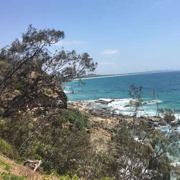 A imagem retrata um litoral rochoso sereno com um oceano azul vibrante, com vegetação exuberante e um céu azul claro, evocando uma sensação de beleza natural e tranquilidade.