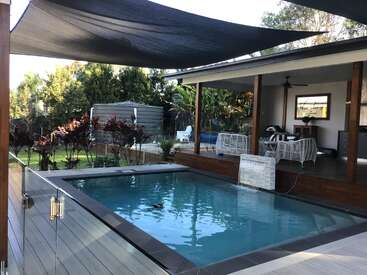 O quintal tem uma piscina retangular com deck, cerca de vidro e vela de sombra. Há um pátio coberto com móveis brancos, cercado por vegetação e plantas exuberantes.