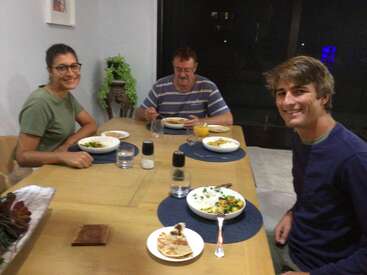 Três pessoas estão sentadas ao redor de uma mesa de jantar de madeira, sorrindo e desfrutando de uma refeição juntas. Há pratos de comida, copos de água e um suco.