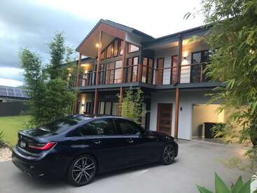 A imagem mostra uma casa moderna de dois andares com um BMW azul escuro estacionado na entrada da garagem, tendo como pano de fundo um céu nublado.