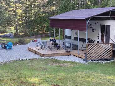 Un acogedor camping cuenta con una caravana con porche cubierto, asientos al aire libre, una persona relajándose con dos perros, rodeada de árboles y la tranquila vegetación de la naturaleza.