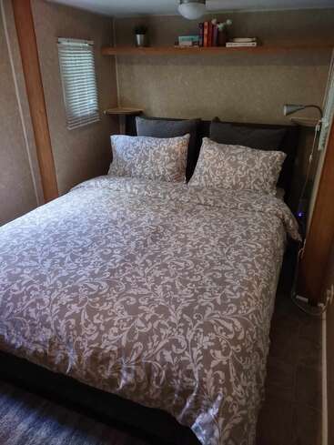 Una acogedora habitación cuenta con una cama perfectamente hecha con un edredón de motivos florales, almohadas, libros en una estantería de madera, persianas, pequeñas lámparas y una iluminación suave.