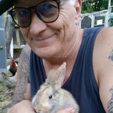 Una persona sonriente con gafas, que lleva un sombrero y una camiseta sin mangas, sostiene con delicadeza un conejo peludo. Entorno al aire libre con árboles, un gallinero y un gallo.