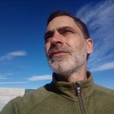 Das Bild zeigt einen Mann mit kurzen dunklen Haaren und einem grünen Pullover vor einem blauen Himmel mit Wolkenfetzen. Er scheint im Freien zu sein.