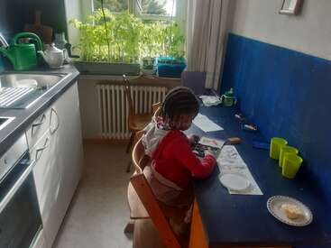 A young child sits at a blue table in a kitchen, coloring pictures. Plants grow on the windowsill, and kitchen items are neatly arranged nearby.