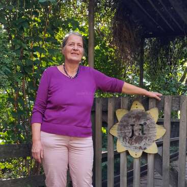 A woman in a purple top and pink pants stands by a wooden gate with a sunflower sign reading “Ja!” surrounded by lush green garden foliage.