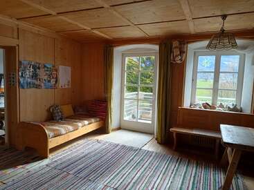 A cozy wooden room with large windows, a daybed, colorful rugs, bench seating, and countryside views. Sunlight brightens the warm, inviting interior. Peaceful, homey atmosphere.