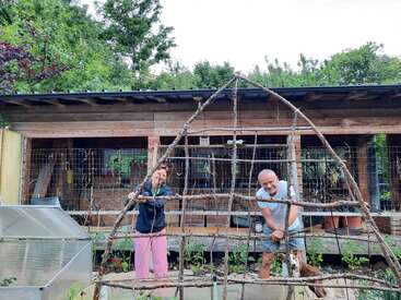 Une femme et un homme se tiennent devant un bâtiment en bois, souriants, tenant une treille en bois naturel dans un jardin, entourés de plantes vertes et d'arbres.