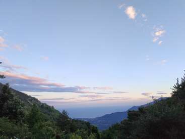 Uma paisagem montanhosa serena com árvores verdes exuberantes em primeiro plano, colinas distantes, mar azul calmo e um vasto céu repleto de nuvens rosa suave.