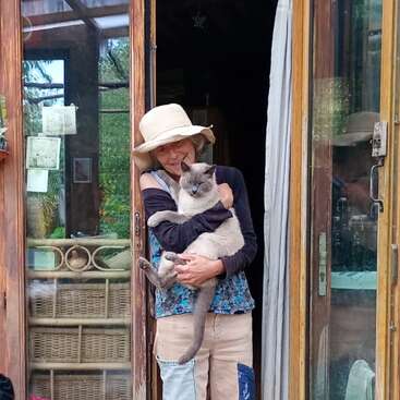 Eine Frau mit Sonnenhut steht in einem rustikalen Hauseingang und hält eine graue Katze, umgeben von Pflanzen, Körben, Holzmöbeln und gemütlicher, heimeliger Dekoration.